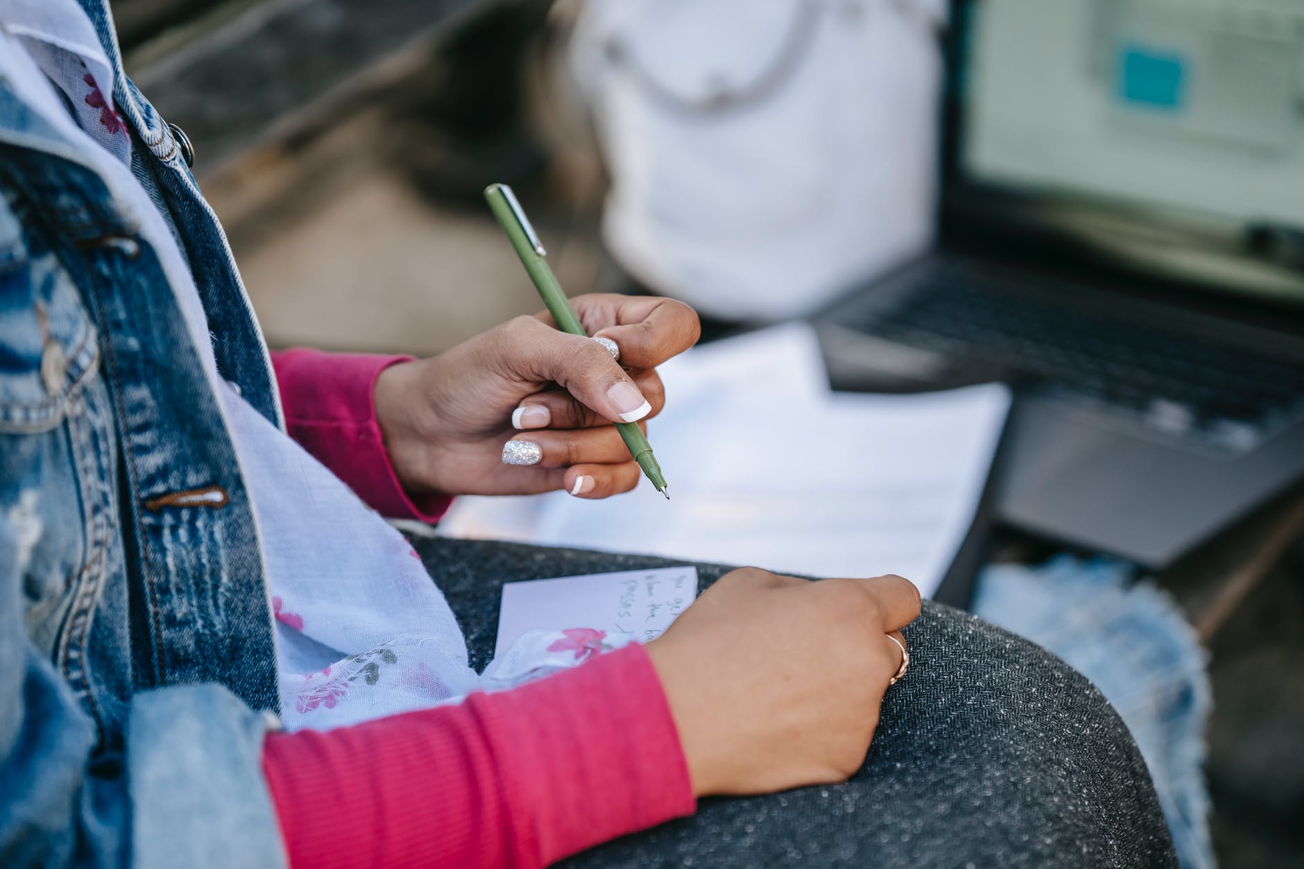 crop female student writing notes while sitting with laptop on bench